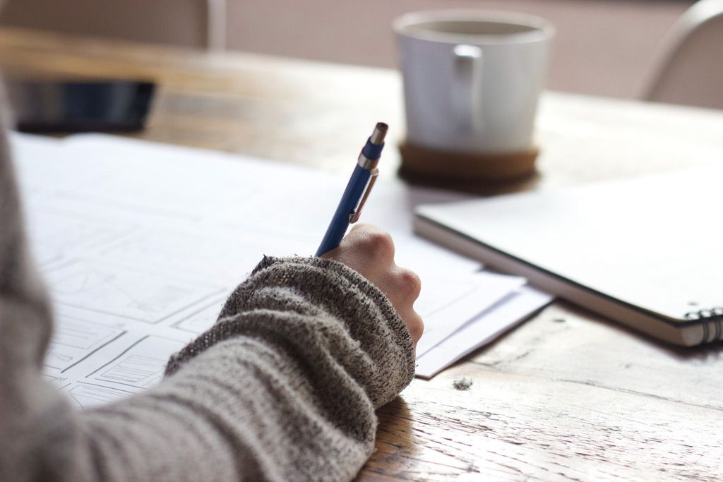person writing using a pen on a desk with a cup of coffee